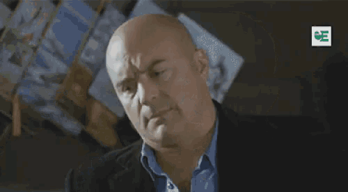 a bald man in a suit and blue shirt is standing in front of a stack of books .