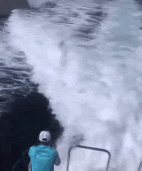a man in a blue shirt is standing on a boat looking at a large wave .