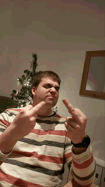 a man in a striped shirt giving the middle finger in front of a christmas tree