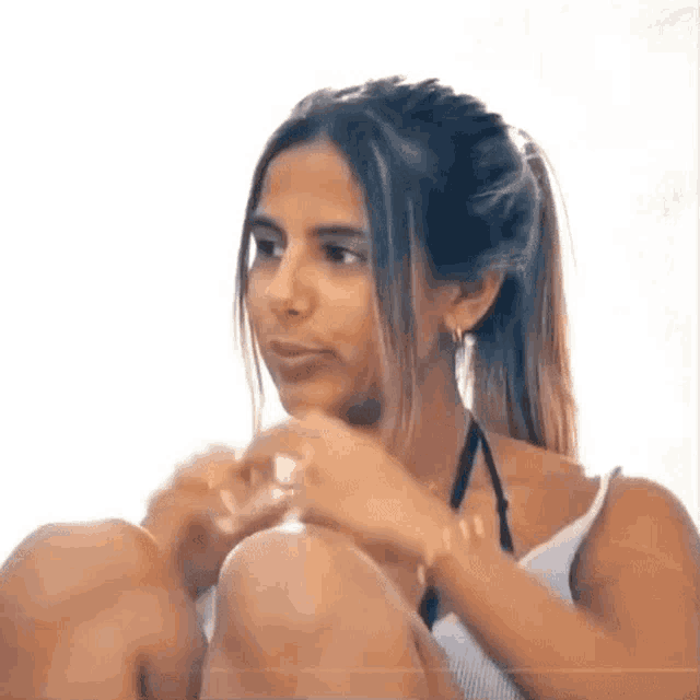 a woman in a white tank top is sitting on the floor with her legs crossed and looking at the camera .