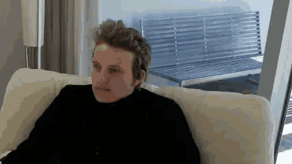a man in a black sweater is sitting on a white couch in front of a window .