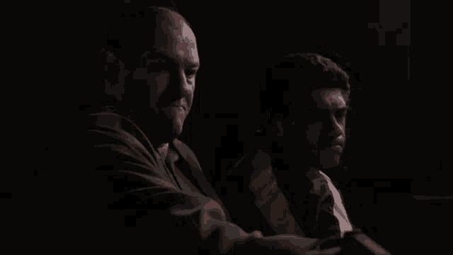 two men are sitting next to each other in a dark room and one of them is looking at the camera .