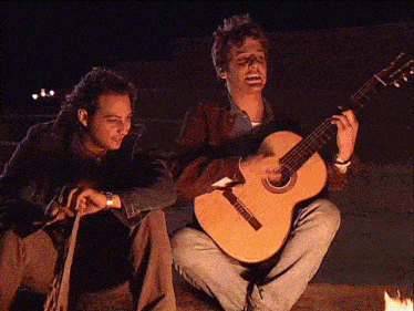 a man sitting next to another man playing a guitar that says ' fender ' on the neck