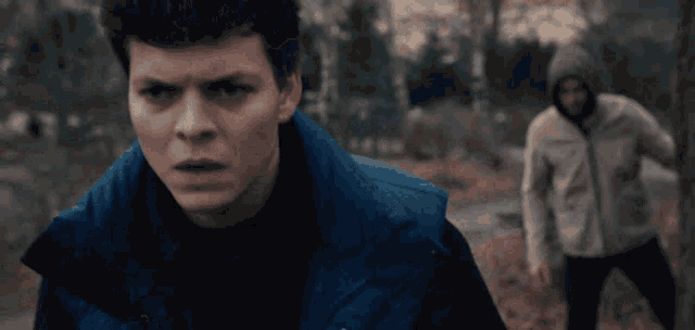 a young man in a blue vest looks at the camera with a serious look on his face