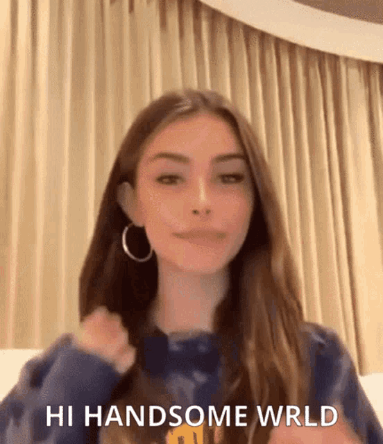 a woman with long hair and hoop earrings is standing in front of a curtain and says `` hi handsome wrld '' .