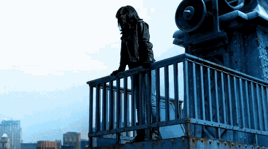 a woman in a black jacket stands on a balcony overlooking the city
