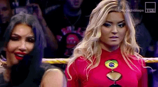 two women are standing next to each other in a wrestling ring . one of the women is wearing a red top .