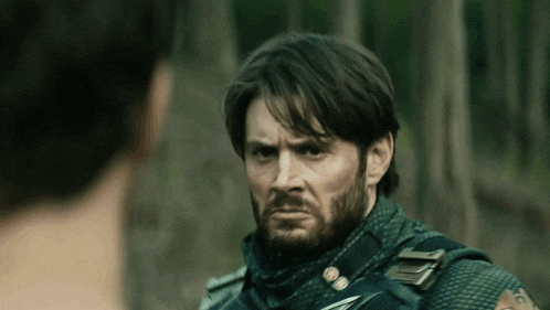 a man with a beard is standing next to another man in the woods .