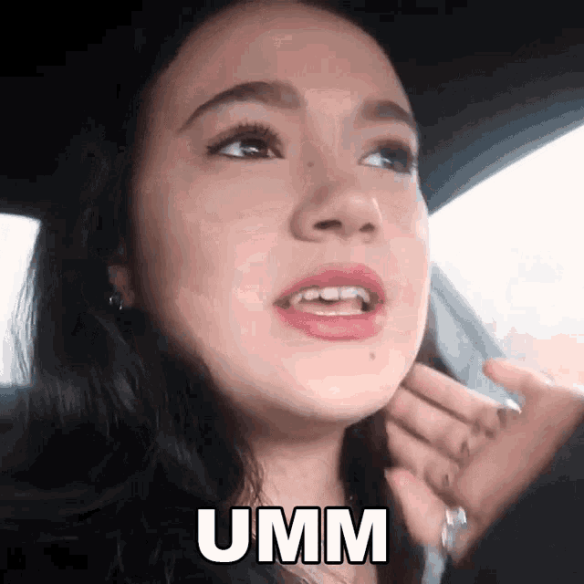 a woman is sitting in a car with her hand on her face and the word umm is on the screen .