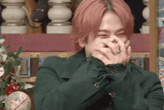 a man with pink hair is covering his mouth with his hands while laughing .
