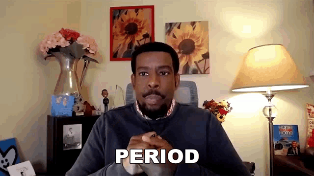 a man with a beard is sitting in front of a lamp and flowers and says period