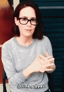 a woman wearing glasses is holding a glass of beer and saying `` drink a beer '' .