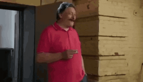 a man in a red shirt with a mustache is standing in front of a brick wall .