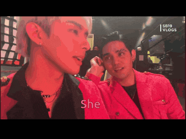 a man in a red jacket is talking to another man in a black shirt with the word she on the bottom