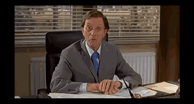 a man in a suit sits at a desk with his hands folded