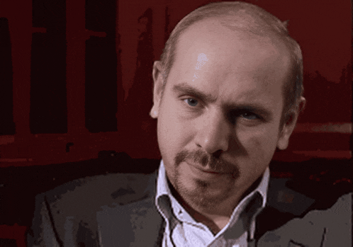a bald man with a beard looks at the camera with a red background