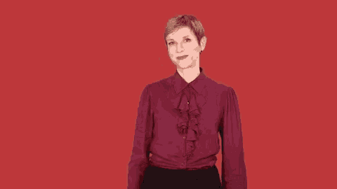 a woman in a burgundy ruffled shirt and black skirt is standing in front of a red background and smiling .