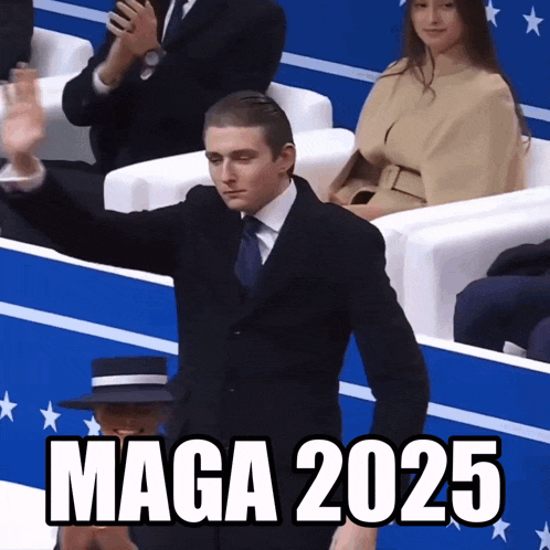 a man in a suit and tie stands in front of a crowd with maga 2025 written on the screen