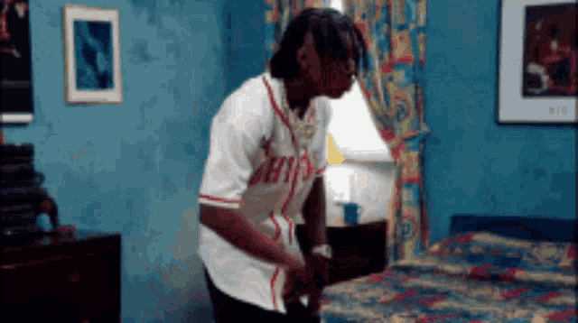 a man in a baseball jersey is standing next to a bed in a room .