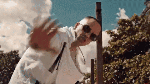 a man wearing sunglasses and a white shirt is waving his hand in front of a tree .