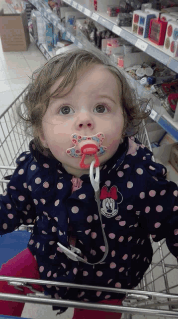 a little girl wearing a minnie mouse jacket is sitting in a shopping cart with a pacifier in her mouth