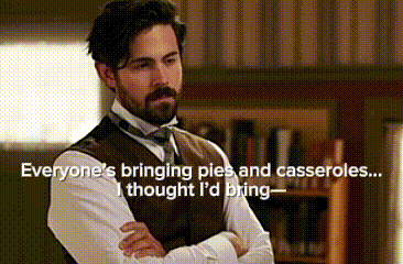 a man with a beard is standing with his arms crossed in front of a bookshelf and a window .