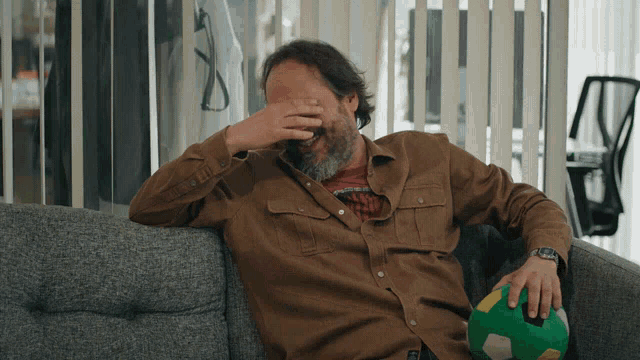 a man sits on a couch covering his face with his hand while holding a soccer ball