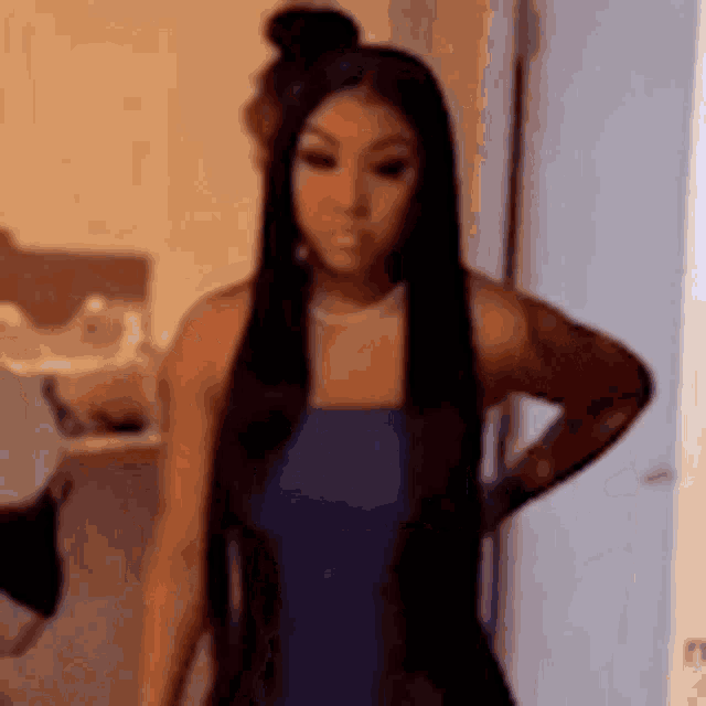 a woman with long black hair is wearing a blue dress and standing in a room with her hands on her hips .