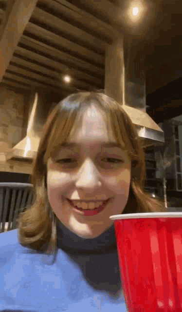 a girl in a blue shirt is holding a red cup and smiling