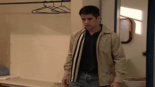 a man in a tan jacket stands in a room with a toilet