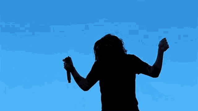 a silhouette of a man holding a microphone with his arms outstretched in front of a blue sky .