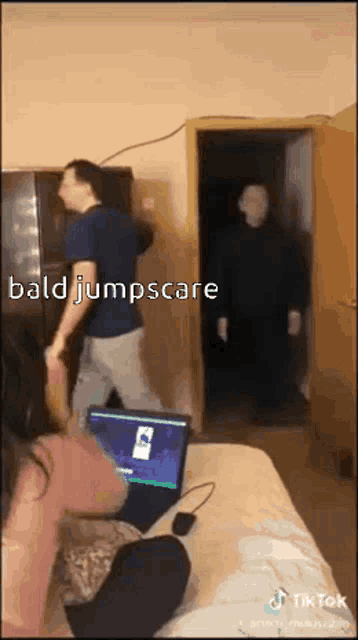 a woman is sitting on a bed with a laptop and a man is standing in the background .