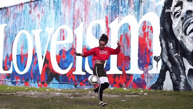 a woman is kicking a soccer ball in front of a wall that has the word love on it