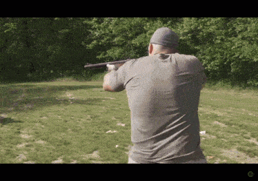 a man in a grey shirt is holding a shotgun in a field with trees in the background