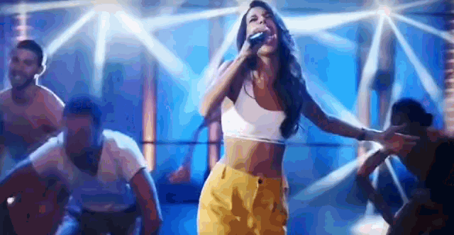 a woman in a crop top and yellow pants is singing into a microphone on a stage in front of a crowd .