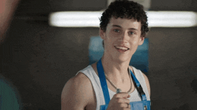 a young man wearing a white tank top and blue overalls smiles