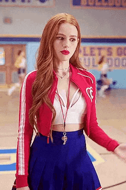 a woman is standing on a basketball court wearing a red jacket , white top , and blue skirt .