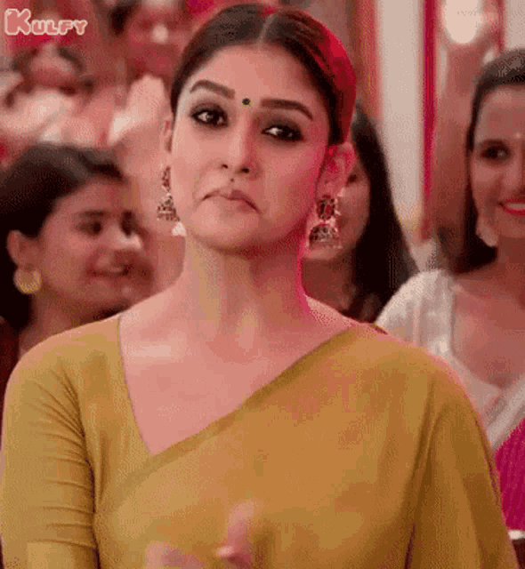 a woman wearing a yellow blouse and earrings is standing in front of a crowd of people .