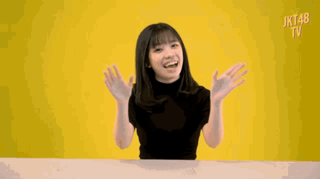a girl sitting at a table with her arms outstretched in front of a yellow background with the words jkt48 tv on it