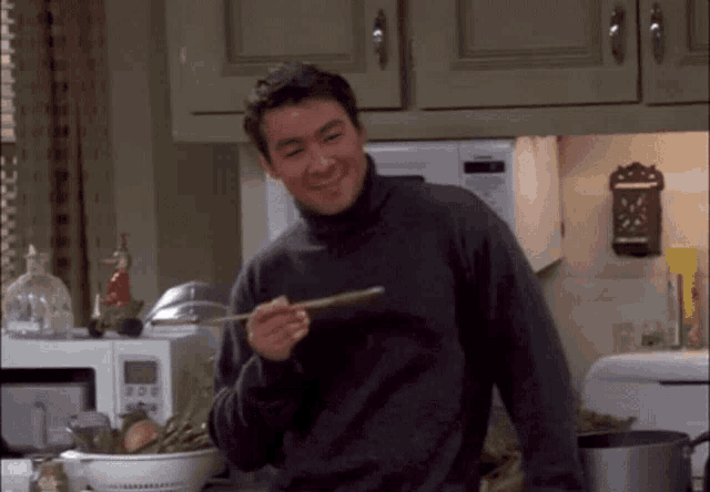 a man in a turtleneck sweater is holding a spoon and smiling in a kitchen