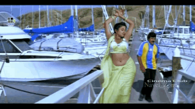a woman in a yellow saree is dancing in front of boats and the words cinema club navel zone are on the bottom of the screen