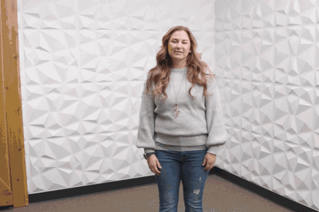 a woman in a grey sweater and jeans stands in a corner of a room