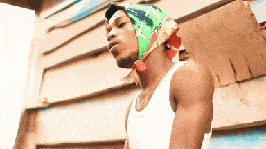 a man wearing a white tank top and a colorful head scarf stands in front of a wooden wall