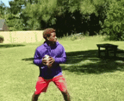 a man in a purple jacket and red shorts holds a football in his hands