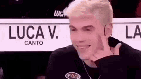 a young man with blonde hair is making a funny face while sitting in front of a sign that says luca v canto .