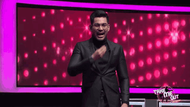 a man in a black suit is standing in front of a screen that says take me out