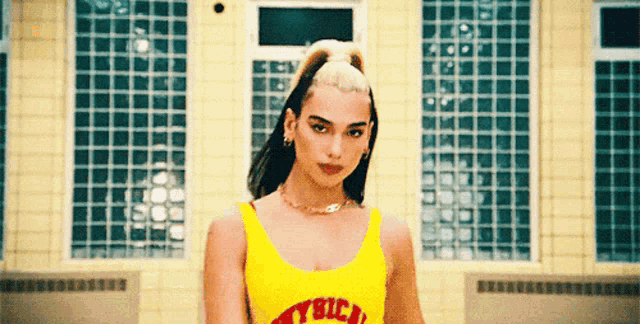a woman in a yellow tank top with the word trick on it is standing in front of a building .