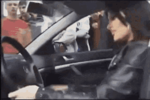 a woman is driving a car while a group of people are standing behind her .