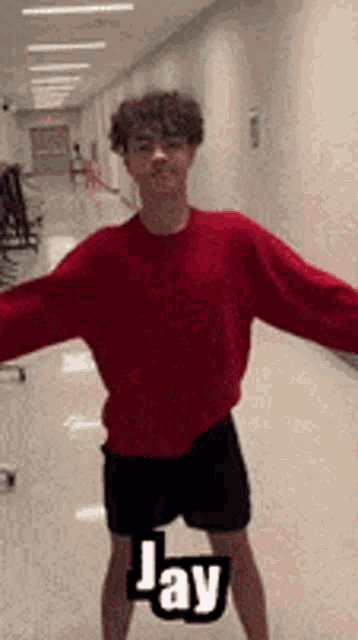a young man wearing a red sweater and black shorts is standing in a hallway with his arms outstretched .