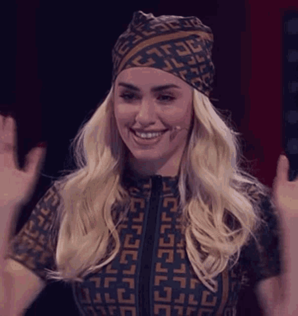 a blonde woman wearing a head scarf and a jumpsuit is smiling and waving .